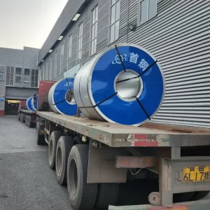 Flatbed trailer loaded with two large blue-edged CRGO electrical steel coils outside a steel warehouse at dusk