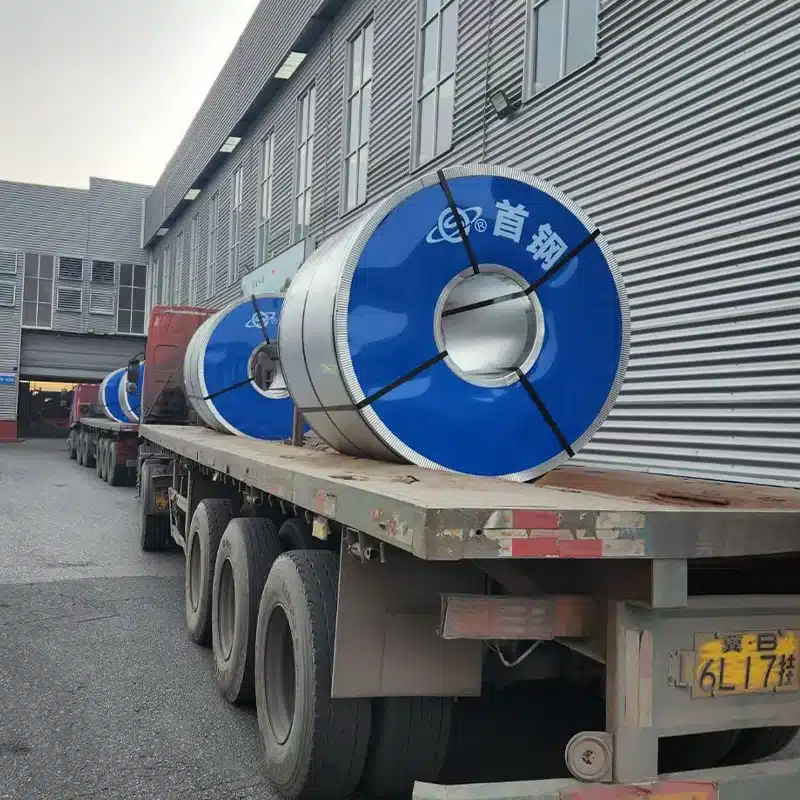 Flatbed trailer loaded with two large blue-edged CRGO electrical steel coils outside a steel warehouse at dusk