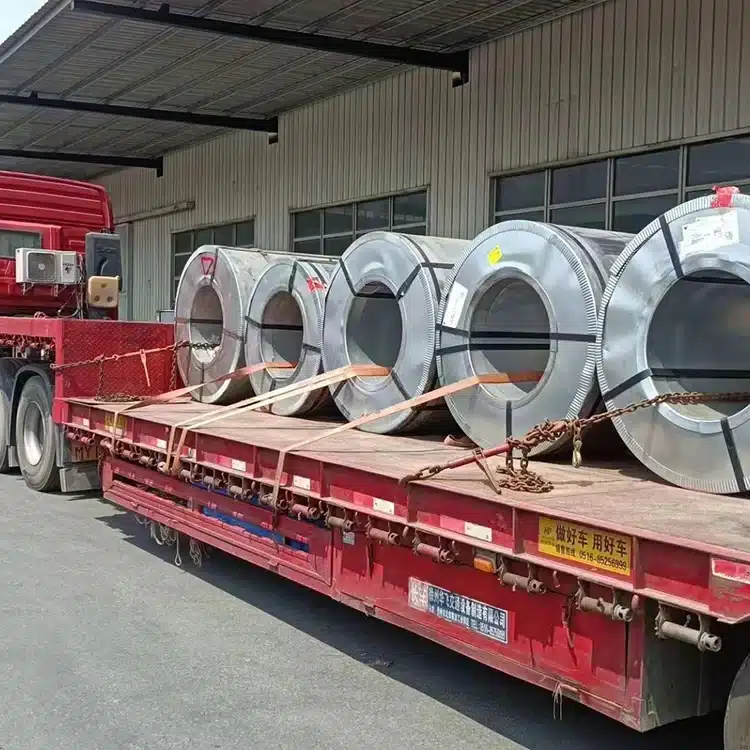 Five silver CRGO electrical steel coils chained down on a red flatbed truck outside a warehouse loading dock