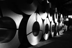 Rows of grain-oriented electrical steel coils stacked in a warehouse, lit from the side