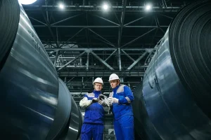 Two steel mill workers reviewing coil test data between rows of silicon steel coils during supplier qualification