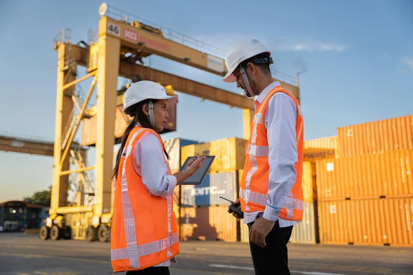 Two logistics workers in hard hats and safety vests reviewing a tablet at a shipping container yard 