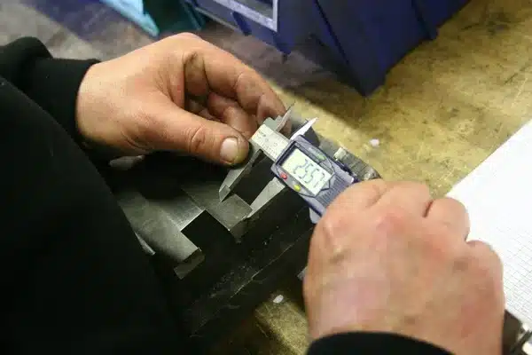 Worker using a digital caliper to measure a steel bar on a workshop bench, representing instrument-based verification