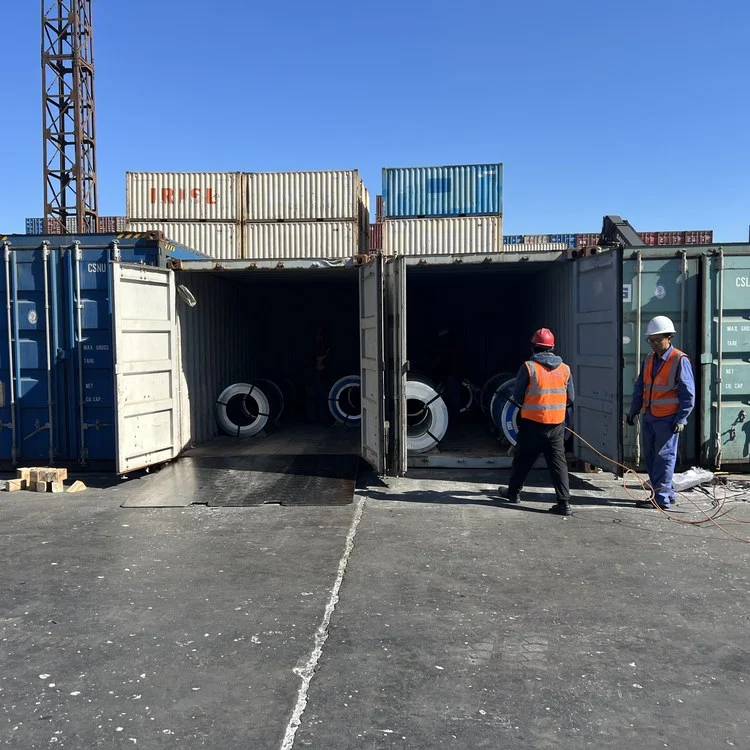 Two workers in hi-vis vests at open shipping containers loaded with grain-oriented steel coils