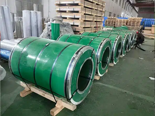 Row of green-wrapped CRGO electrical steel coils on wooden cradles staged in a warehouse, with additional wrapped coils and packaging materials in the background