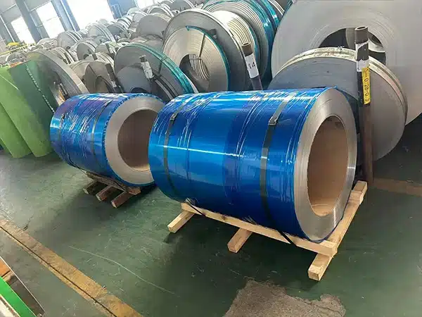 Warehouse rack storage of electrical steel coils, with two blue-wrapped coils on wooden pallets in the foreground and additional coils in teal wrap and bare finish stacked behind