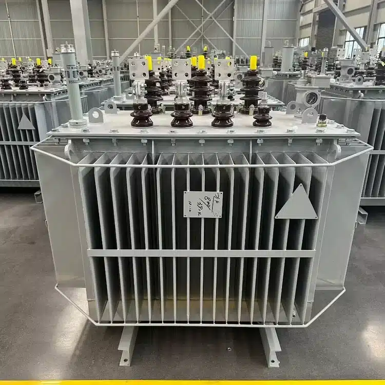 A three-phase oil-immersed distribution transformer core and tank assembly on a factory floor, bushings and conservator fittings mounted on top