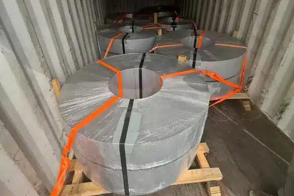 Six slit coils of 0.10mm GT-100 high frequency reactor core steel laid eye-to-sky on wooden pallets inside a shipping container, secured with orange ratchet straps