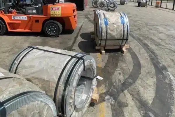 Wrapped ultra-thin electrical steel coils staged on wooden cradles in an outdoor yard with a forklift, ahead of container loading